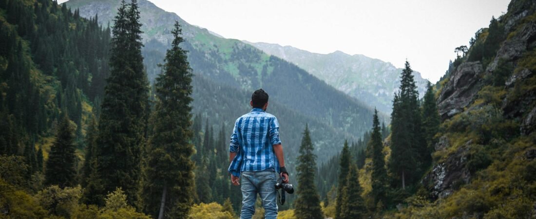 solo traveler photographer man in blue and white checked quarter sleeved shirt holding black camera standing on rock front of nature landscape